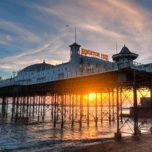 Un grande molo con edifici bianchi si estende sull'oceano da una spiaggia di ciottoli durante un tramonto dorato.