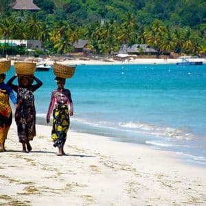 Drei Frauen in farbenfroher Kleidung gehen an einem Sandstrand entlang, jede mit einem Weidenkorb auf dem Kopf, neben türkisfarbenem Wasser.