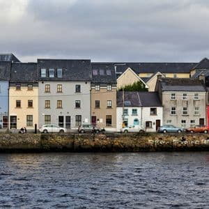 Une rangée de maisons colorées avec des voitures garées se dresse sur un quai en pierre au bord de l'eau sous un ciel nuageux.
