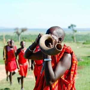 Una persona con atuendo tradicional rojo sopla un gran cuerno mientras un grupo camina por una llanura verde en el fondo.