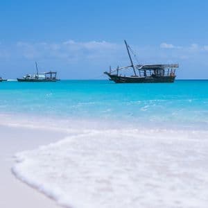 Three traditional wooden boats anchored in turquoise water, seen from a white sand beach under a clear blue sky.