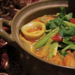 Un primo piano di un curry di mare con anelli di calamaro, sedano e coriandolo, servito in una ciotola di ceramica rustica su una tovaglia fantasia.