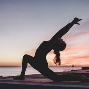 Silhouette di una persona che pratica yoga in posizione affondo su un tappetino in riva al mare al tramonto.