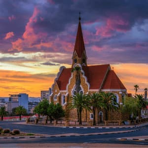 Una chiesa in pietra con tetto rosso e campanile su uno spartitraffico cittadino sotto un cielo drammatico al tramonto viola e arancione.
