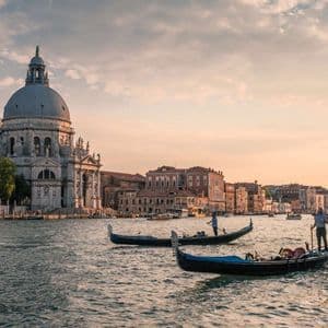 Gondole galleggiano su un canale di fronte a una grande basilica a cupola e a edifici storici al tramonto.