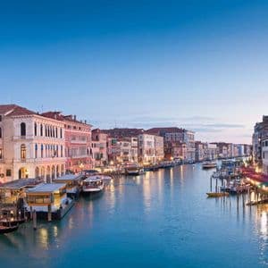 Illuminated historic buildings line a wide canal filled with boats and vaporettos under a clear blue sky at dusk.