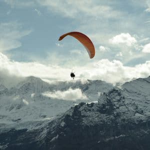 Deux personnes en parapente biplace avec une aile orange s'envolent au-dessus d'une vaste chaîne de montagnes enneigée sous un ciel nuageux.