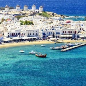 Una vista dall'alto di una cittadina costiera con edifici bianchi, una piccola spiaggia e mulini a vento affacciati su un mare turchese con barche.