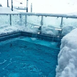 An outdoor hot tub with two waterfall jets flowing into the blue water, surrounded by deep snow at a mountain resort.