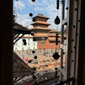Ein Blick aus einem Fenster auf einen belebten öffentlichen Platz mit Marktständen, Menschen und einem traditionellen Pagodentempel im Hintergrund.