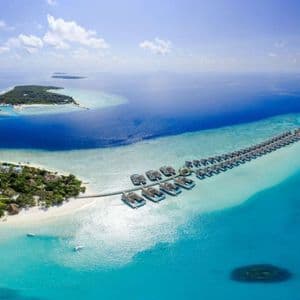 Vista aérea de un resort en isla tropical con dos largas filas de villas sobre el agua que se extienden en un mar turquesa bajo un cielo nublado.