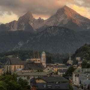 Ein Alpendorf mit markanten Kirchtürmen liegt bei Sonnenuntergang am Fuße hoch aufragender, felsiger Berge.