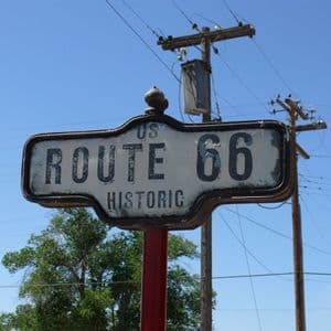 Primer plano de un cartel desgastado de la Ruta Histórica 66 de EE. UU. en un poste rojo, con un cielo azul y postes de servicios de fondo.