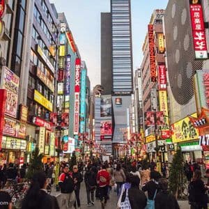 Una folla di persone cammina lungo una strada cittadina fiancheggiata da edifici coperti da colorate insegne al neon, con una grande testa di Godzilla visibile.