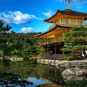 Un tempio dorato a più livelli si riflette in un lago calmo, circondato da alberi verdi sotto un cielo blu.