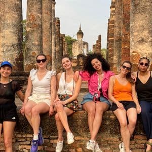A WeRoad group trip of six women sitting on a brick wall, posing for a photo in front of ancient temple ruins with a Buddha statue.