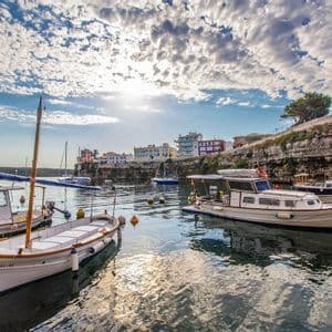 Mehrere Boote liegen in einem ruhigen Hafen neben einer Klippe mit Gebäuden unter einem teilweise bewölkten Himmel.