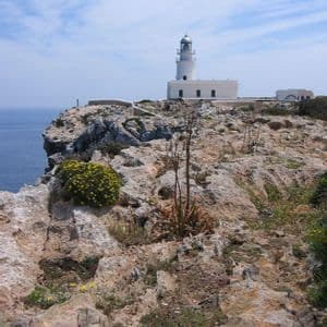 Ein weißer Leuchtturm thront auf einer felsigen Klippe mit Blick auf das blaue Meer unter einem teils bewölkten Himmel.