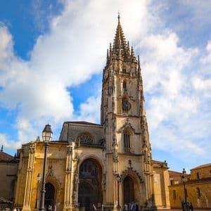 Una ornamentada catedral gótica con una alta aguja se alza en una plaza de piedra bajo un cielo azul con nubes blancas.