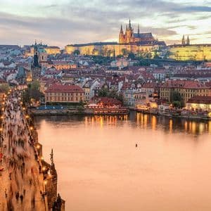 An illuminated stone bridge crowded with people crosses a river, leading to a historic cityscape with a castle on a hill at dusk.