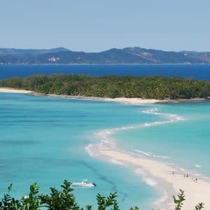Veduta aerea di una sinuosa lingua di sabbia bianca che porta a un'isola tropicale lussureggiante, con alcune persone che vi camminano e una barca nel mare turchese.