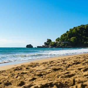 Una vista dal basso di una spiaggia di sabbia con onde dolci, un mare turchese e una costa verde e alberata sotto un cielo azzurro e sereno.