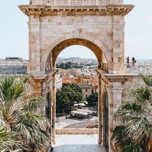 Una ciudad con edificios de techos de terracota se ve a través de un gran arco de piedra, flanqueado por palmeras en primer plano.