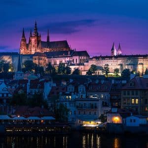 Un castello e una cattedrale illuminati su una collina che domina gli edifici della città e un fiume al crepuscolo sotto un cielo viola