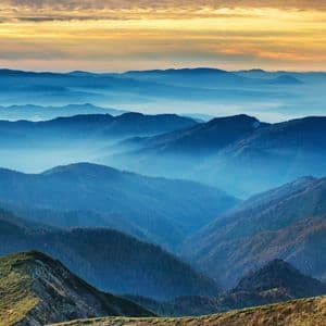 Catene montuose blu si estendono in lontananza, con valli avvolte nella nebbia sotto un cielo dorato al tramonto.
