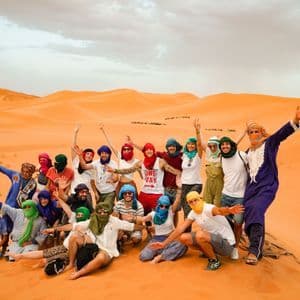 A large WeRoad group trip wearing colorful headscarves and turbans poses for a group photo on a sand dune in the desert.