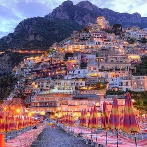 Edifici illuminati su una scogliera che si affaccia su una spiaggia di ciottoli con file di sedie e ombrelloni al tramonto.