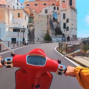 Paseo en primera persona en scooter roja por una carretera sinuosa entre edificios históricos en un pueblo costero soleado.