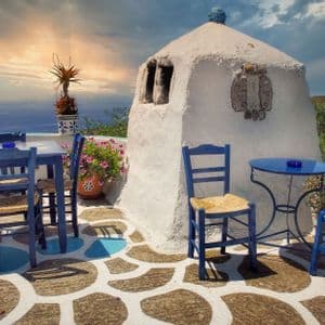 Tavoli e sedie blu su un patio in pietra affacciato sul mare, accanto a un edificio imbiancato a calce al tramonto.