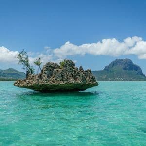 Une grande formation rocheuse avec de petits arbres se trouve dans une eau turquoise claire, avec des montagnes en arrière-plan.