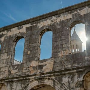 Vue en contre-plongée d'une ruine en pierre patinée aux ouvertures voûtées, avec un clocher visible sur un ciel bleu ensoleillé.