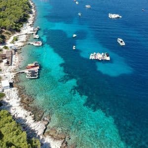 Una vista aerea di una costa rocciosa e alberata con numerose imbarcazioni che galleggiano in acque cristalline color turchese e blu profondo.