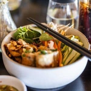 Nahaufnahme einer weißen Schale mit asiatischem Nudelsalat und Essstäbchen, auf einem dunklen Tisch mit einem Glas Limo im Hintergrund.