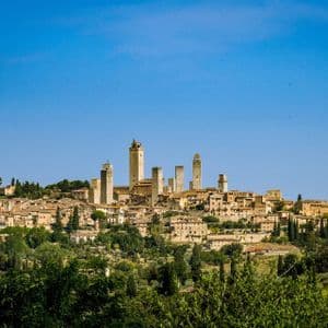 Una città storica con molteplici torri in pietra sorge su una collina verde lussureggiante, circondata da alberi sotto un cielo azzurro limpido.