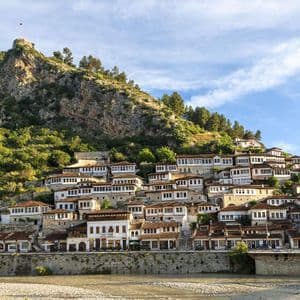 Casas de paredes blancas y tejados de tejas construidas en una exuberante ladera verde, con una fortaleza en la cima y un río debajo.
