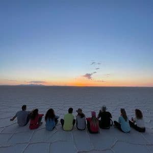 Eine WeRoad Gruppenreise von hinten gesehen, sitzend auf einer weiten Salzwüste, den Sonnenuntergang am Horizont betrachtend.