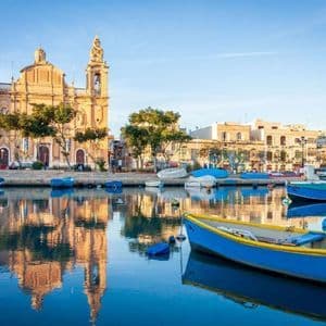 Una grande chiesa in pietra con due campanili si riflette nelle acque calme di un porto pieno di piccole barche colorate.