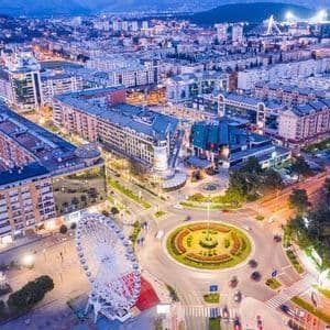 Una vista aerea di una città illuminata al crepuscolo, con una grande ruota panoramica e una rotatoria stradale in primo piano.