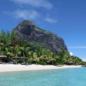 Ein tropischer weißer Sandstrand mit Palmen und Liegestühlen liegt an türkisfarbenem Wasser vor einem großen felsigen Berg.