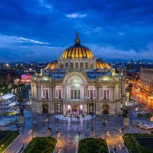 Un sontuoso palazzo con cupola dorata, splendidamente illuminato contro lo skyline cittadino al tramonto, con giardini in primo piano.