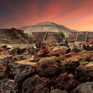 Ancient stone pyramids stand behind a foreground of rough volcanic rocks under a vibrant sunset.