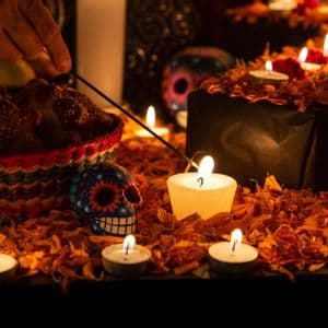 Una mano accende un bastoncino d'incenso su un altare illuminato da candele con teschi di zucchero, pan de muerto e petali di calendula per il Día de los Muertos.