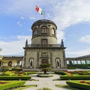 The Mexican flag flies atop a stone tower in a courtyard with manicured gardens, surrounded by colonnades under a blue sky.