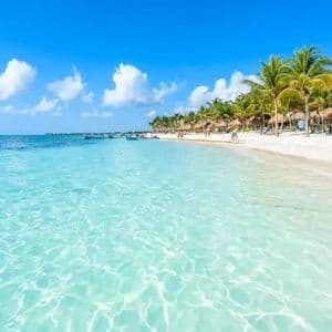 Clear turquoise water meets a white sand beach lined with palm trees and thatched umbrellas under a blue sky.