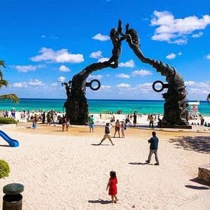 A large sculpture frames a busy tropical beach with people, a playground, and turquoise water under a blue sky.