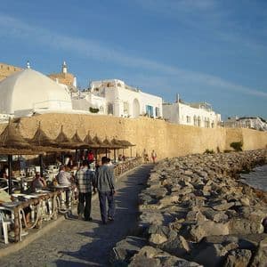 Persone passeggiano e si siedono a un caffè all'aperto con ombrelloni di paglia, lungo una passeggiata in pietra sul mare, con una muraglia fortificata ed edifici bianchi sullo sfondo.